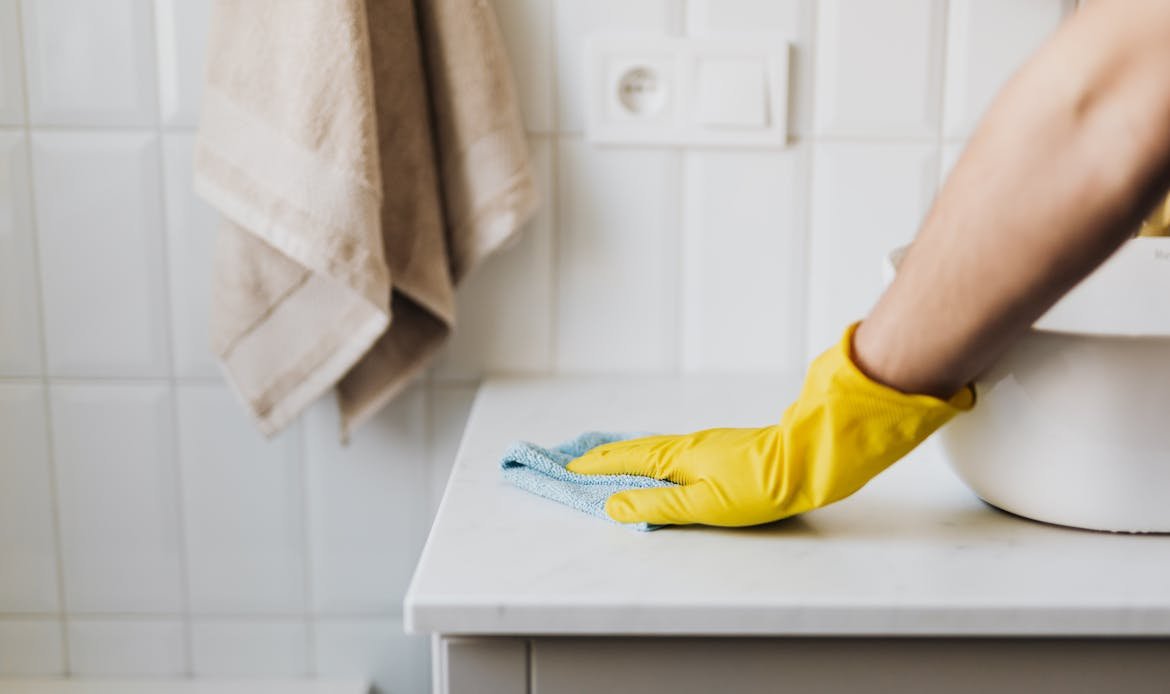 Close-up of a person wearing yellow gloves wiping a bathroom counter with a cloth.